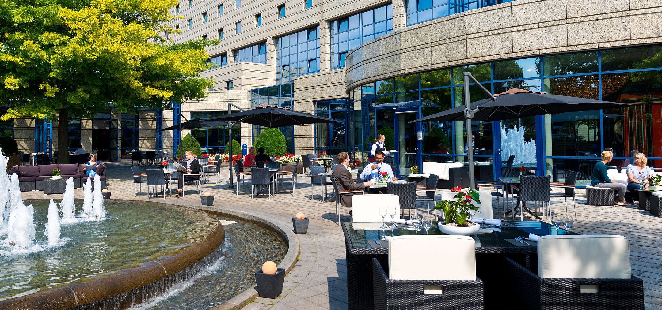 Terrasse des Maritim Hotels Bonn mit Sitzbereichen, Springbrunnen und Gästen, die im Freien speisen und entspannen.
