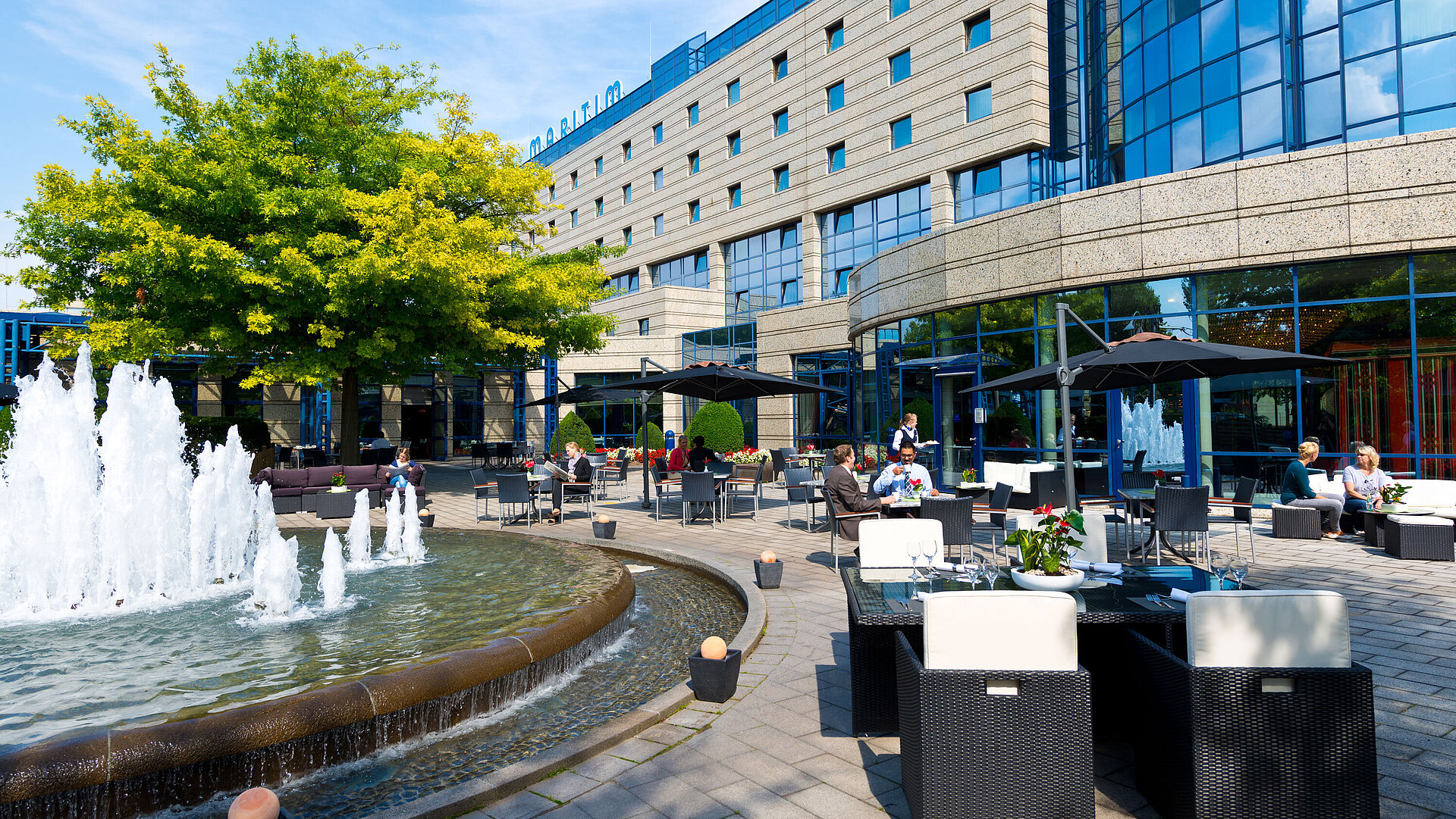 Terrasse des Maritim Hotels Bonn mit Sitzbereichen, Springbrunnen und Gästen, die im Freien speisen und entspannen.