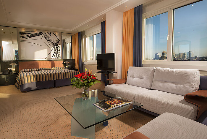 Kuckei-Suite im Maritim proArte Hotel Berlin mit Sofa, Glas-Couchtisch und großem Fenster mit Blick auf die Berliner Skyline.