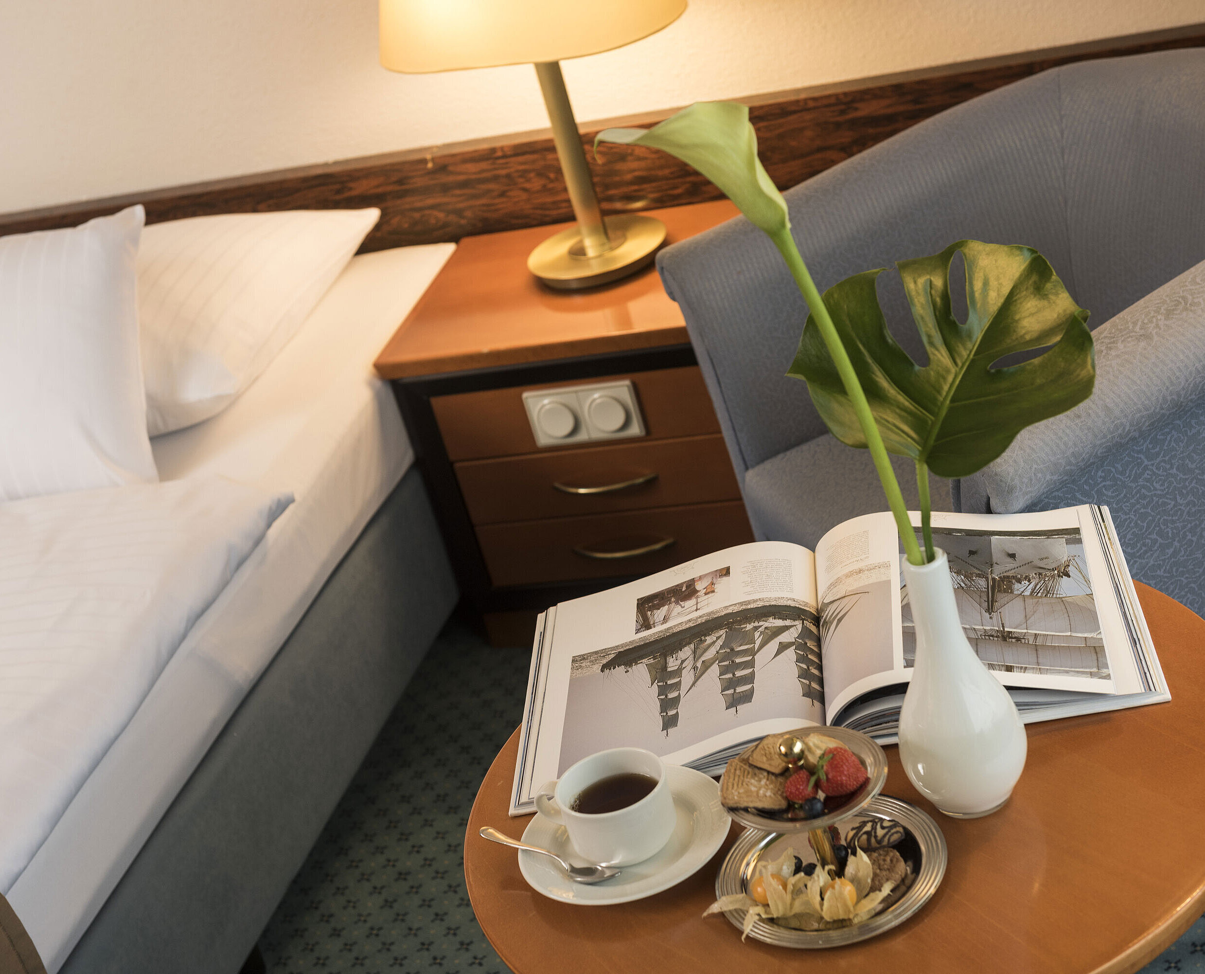 Zimmerdetail Details eines Zimmers im Maritim Hotel Kiel: Beistelltisch mit Vase, Buch und Snacks neben einem Bett und Sessel.