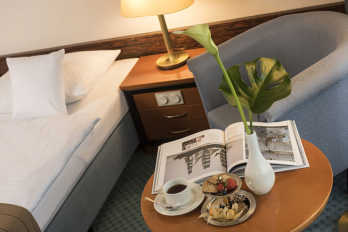 Details eines Zimmers im Maritim Hotel Kiel: Beistelltisch mit Vase, Buch und Snacks neben einem Bett und Sessel.