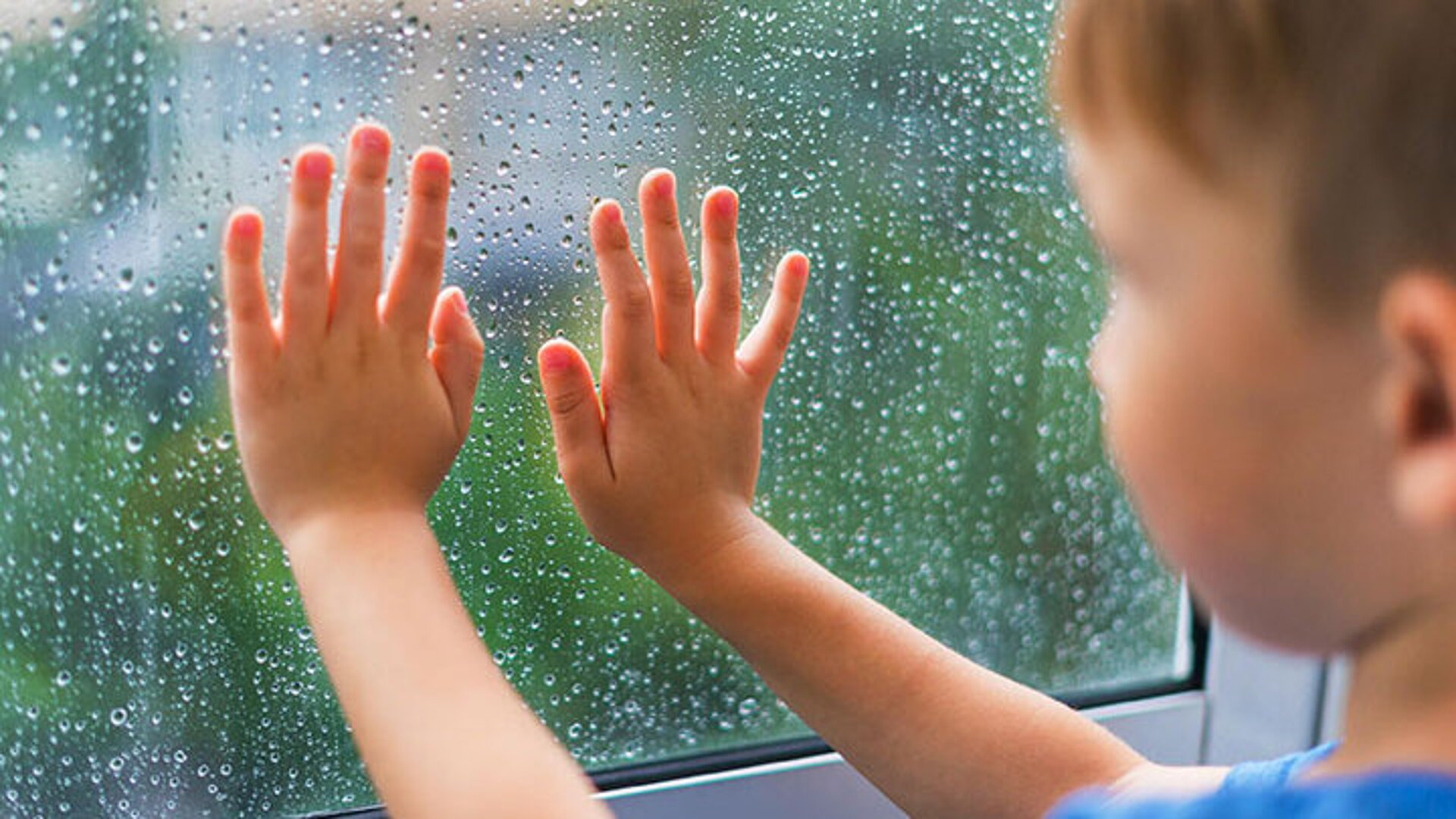 Kind legt beide Hände an eine regennasse Fensterscheibe und schaut nach draußen.
