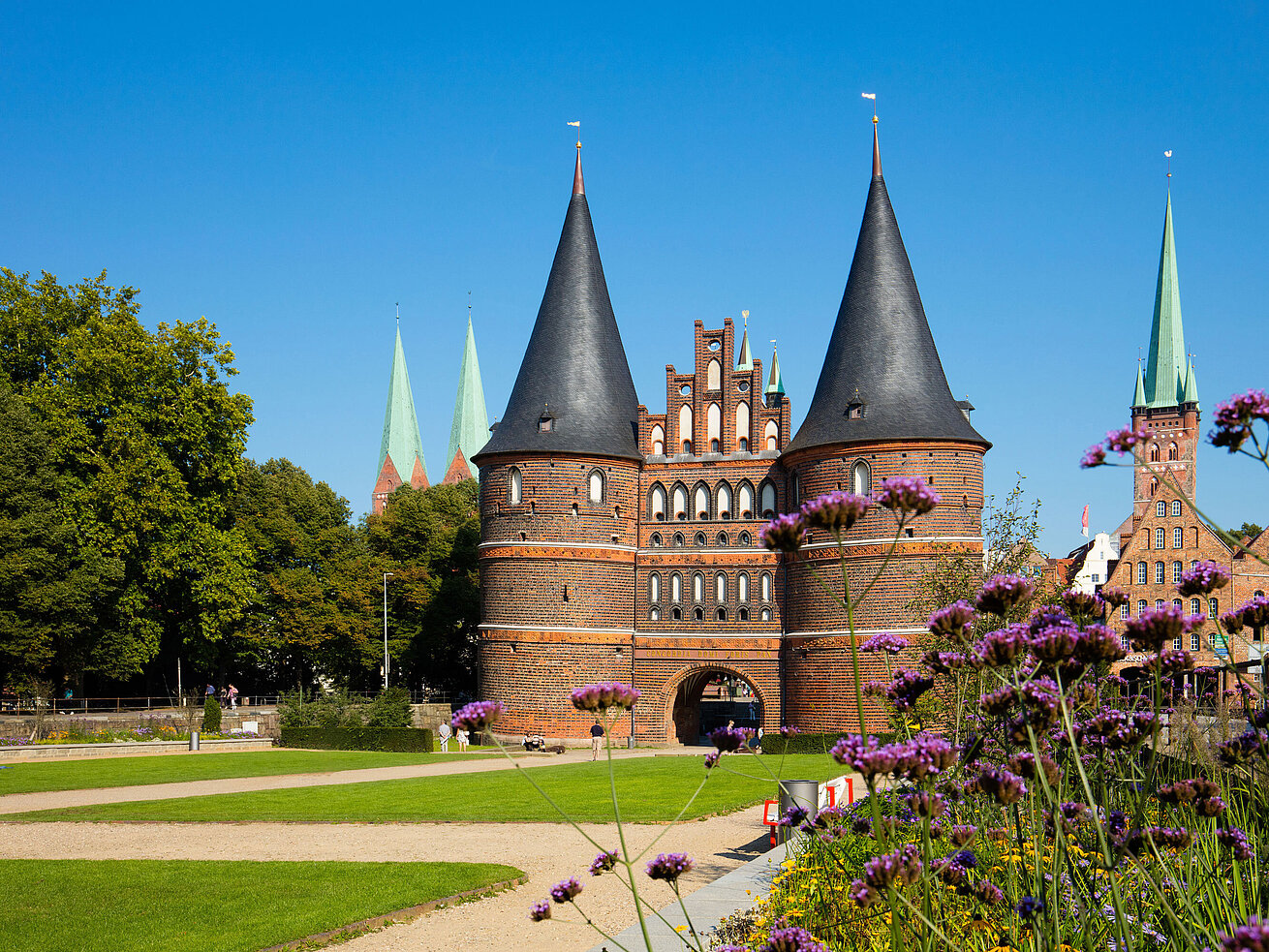 Holstentor in Lübeck mit blühenden Blumen im Vordergrund und blauem Himmel.