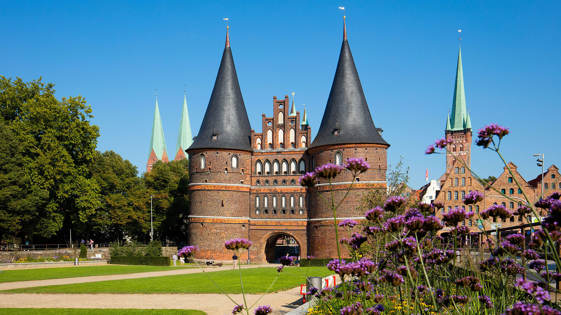 Holstentor in Lübeck mit blühenden Blumen im Vordergrund und blauem Himmel.