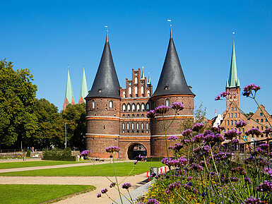 Holstentor in Lübeck mit blühenden Blumen im Vordergrund und blauem Himmel.