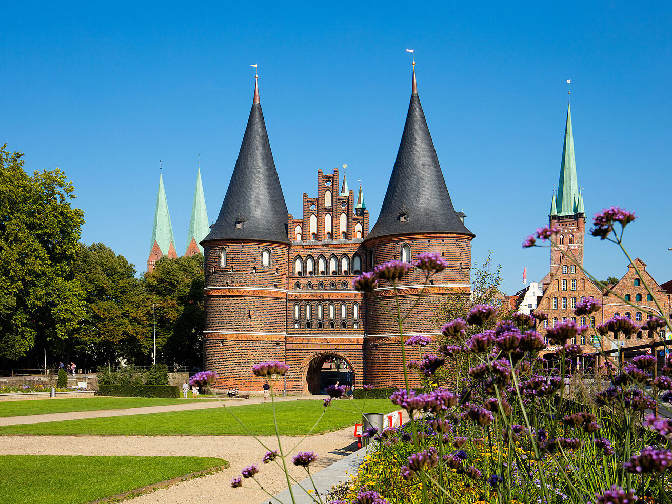 Holstentor in Lübeck mit blühenden Blumen im Vordergrund und blauem Himmel.