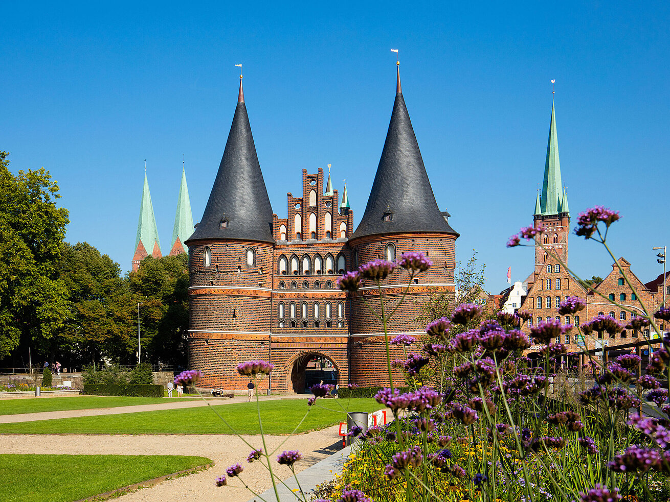 Holstentor in Lübeck mit blühenden Blumen im Vordergrund und blauem Himmel.