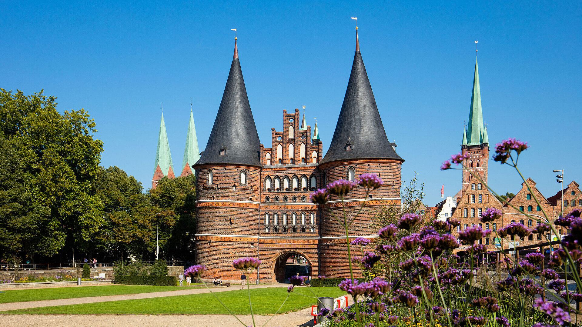 Holstentor in Lübeck mit blühenden Blumen im Vordergrund und blauem Himmel.