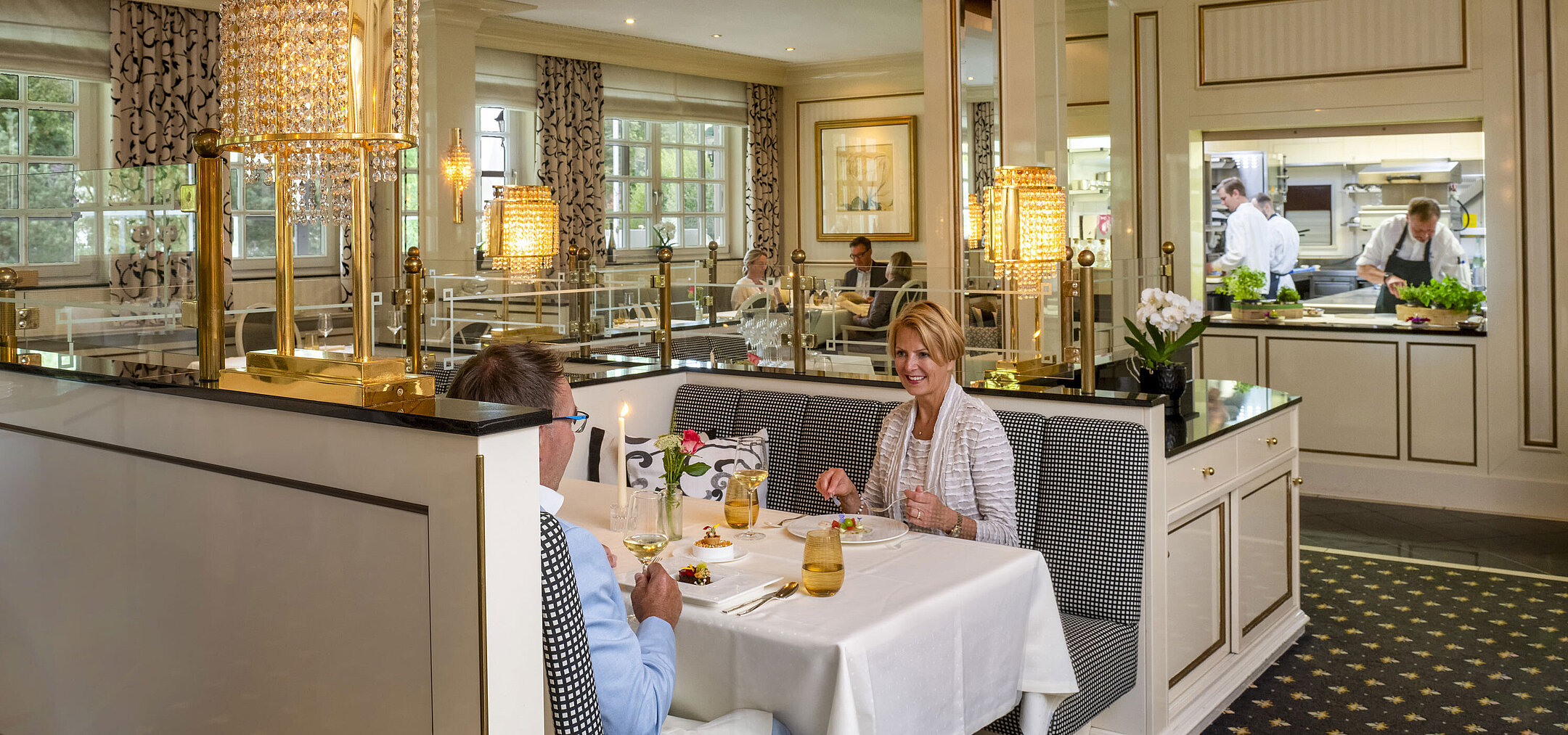 Paar genießt ein feines Dinner im luxuriösen Restaurant Orangerie des Maritim Timmendorfer Strand mit Blick in die Küche.