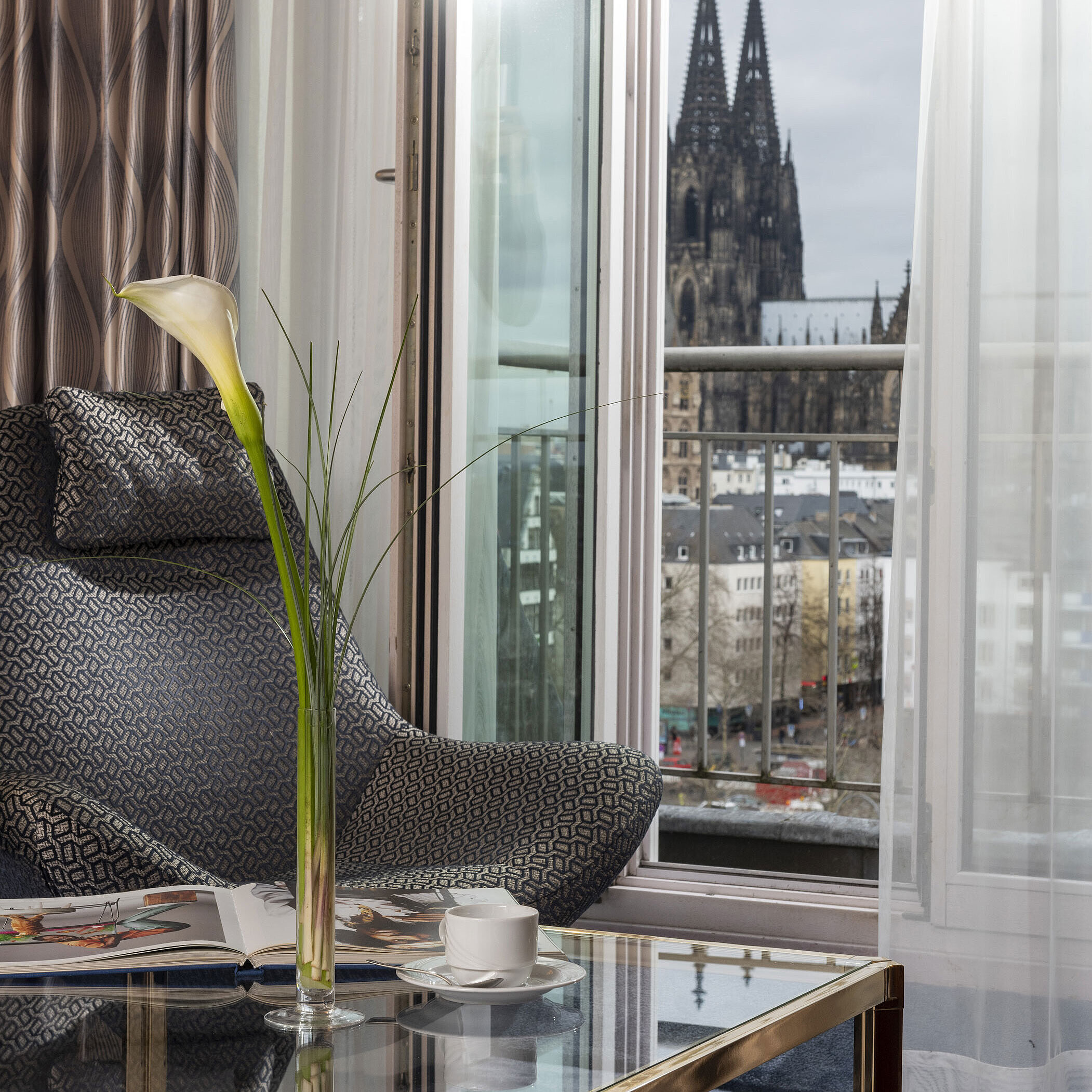 Junior Suite Junior Suite im Maritim Hotel Köln mit Balkonblick auf den Kölner Dom, modernem Sessel und Couchtisch mit Dekoration.