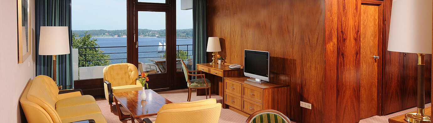 Junior Suite Junior Suite im Maritim Hotel Kiel, gemütliche Sitzecke, Schreibtisch, TV und Balkon mit Blick auf das Wasser.