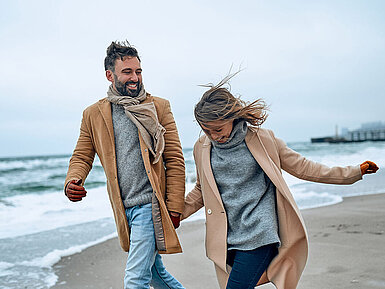 © Valerii Apetroaiei - AdobeStock.com Paar spaziert an einem windigen Tag in warmer Kleidung am Strand entlang.