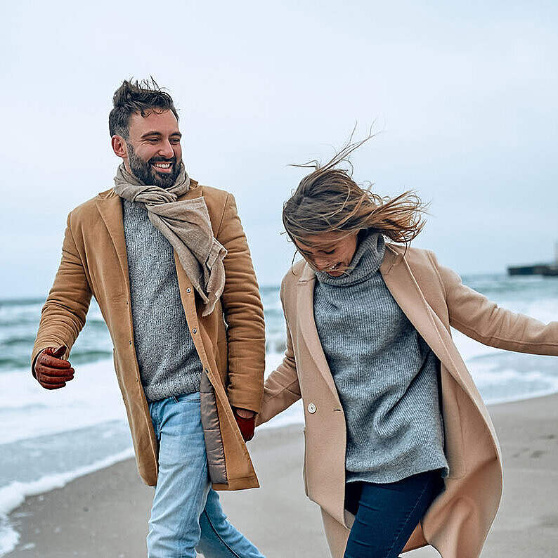 Paar spaziert an einem windigen Tag in warmer Kleidung am Strand entlang.