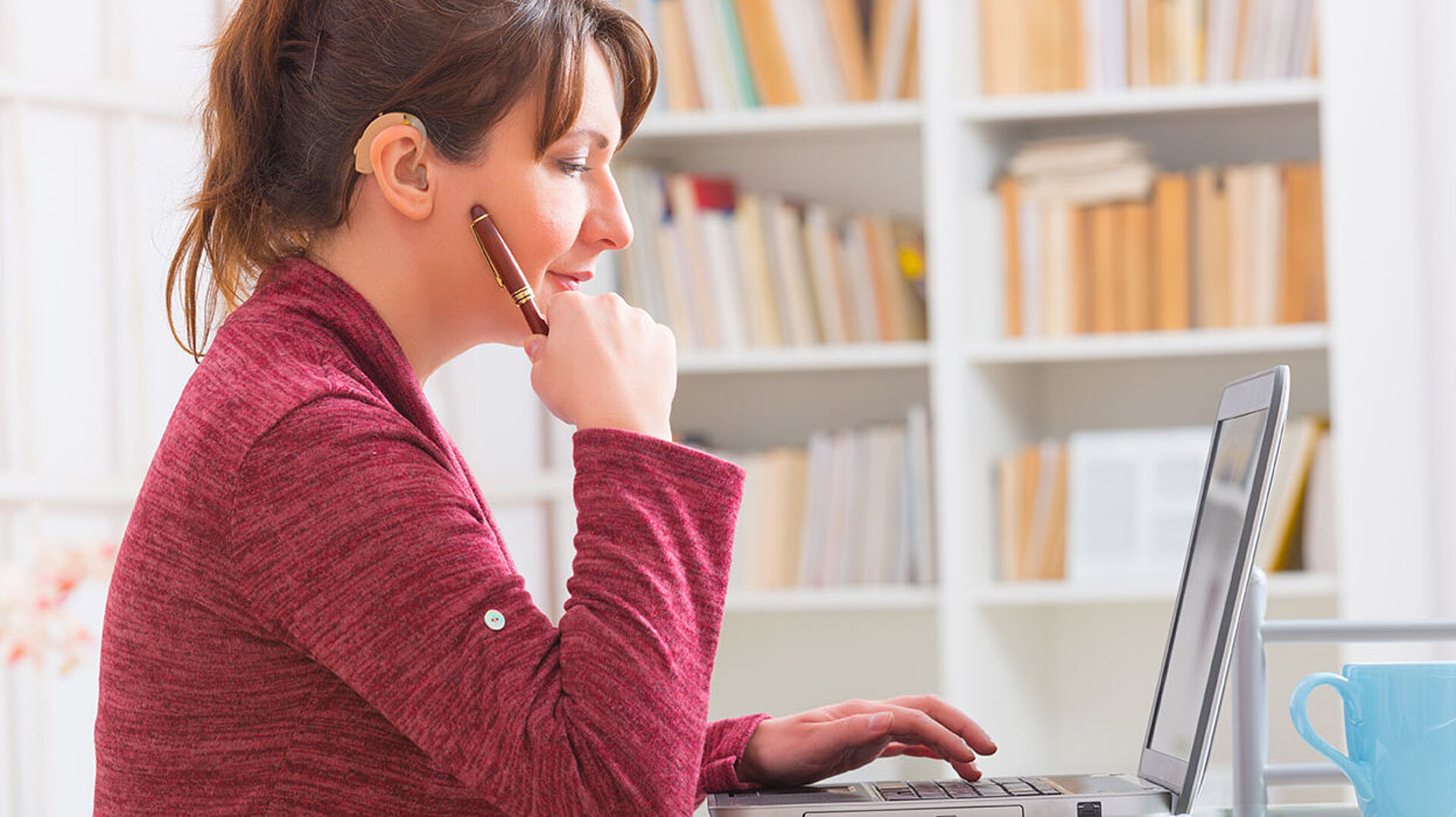 © Monika Wisniewska - AdobeStock.com Frau mit Hörgerät arbeitet konzentriert am Laptop im Homeoffice vor einem Bücherregal.