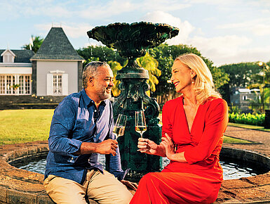 Paar genießt ein Glas Wein am Brunnen im gepflegten Garten bei Abendstimmung