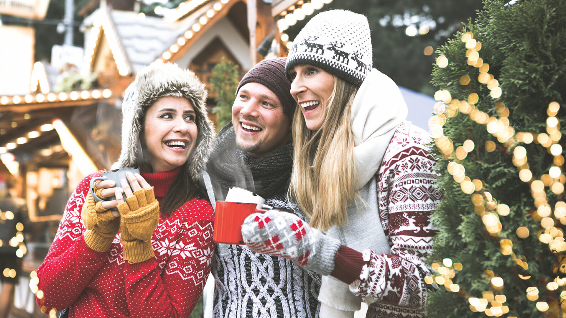 Drei lachende Freunde in Winterkleidung mit heißen Getränken auf einem Weihnachtsmarkt