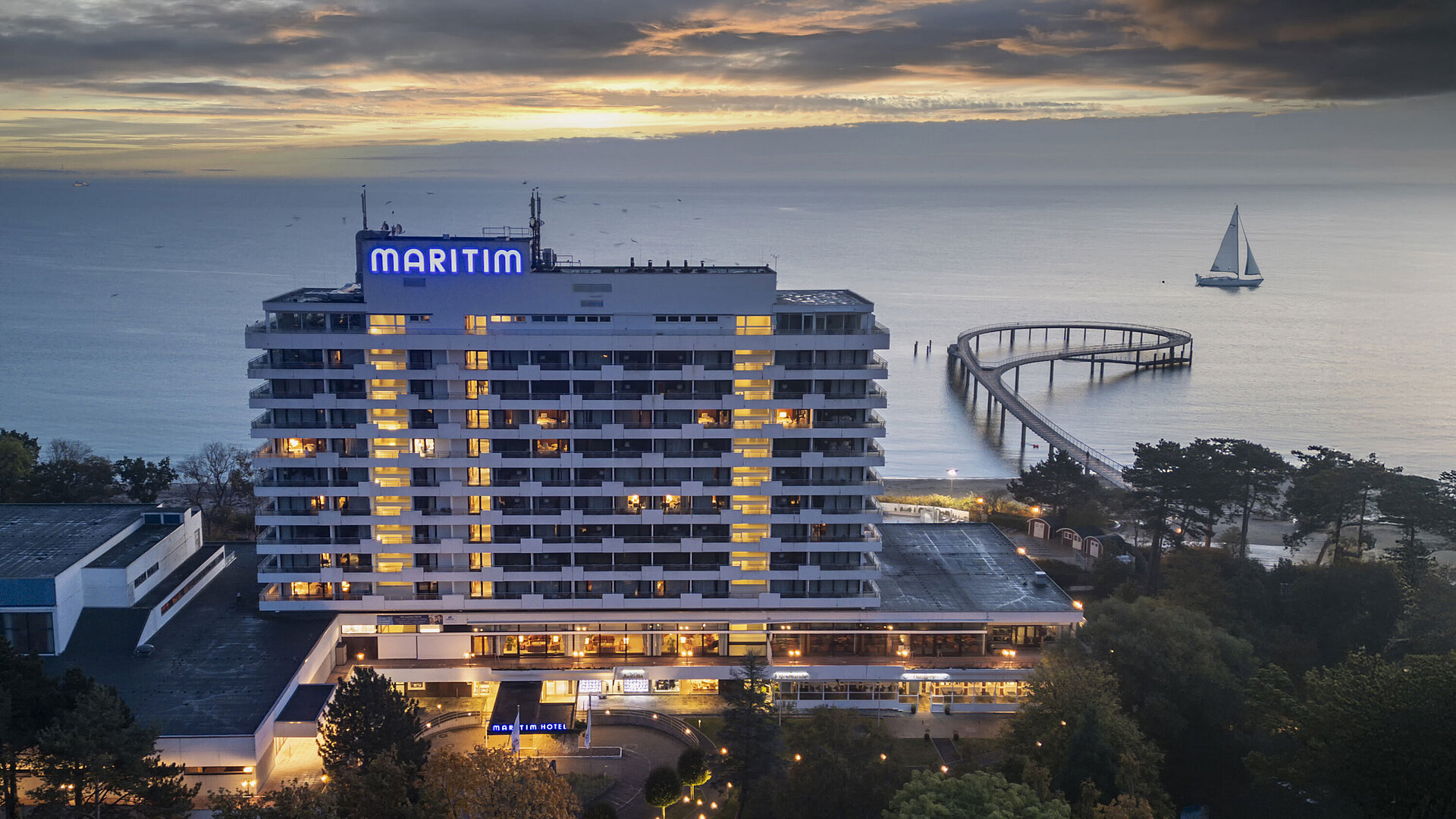 Abendaufnahme des Maritim Seehotels mit beleuchteter Seebrücke und einem Segelboot im Hintergrund bei Sonnenuntergang.