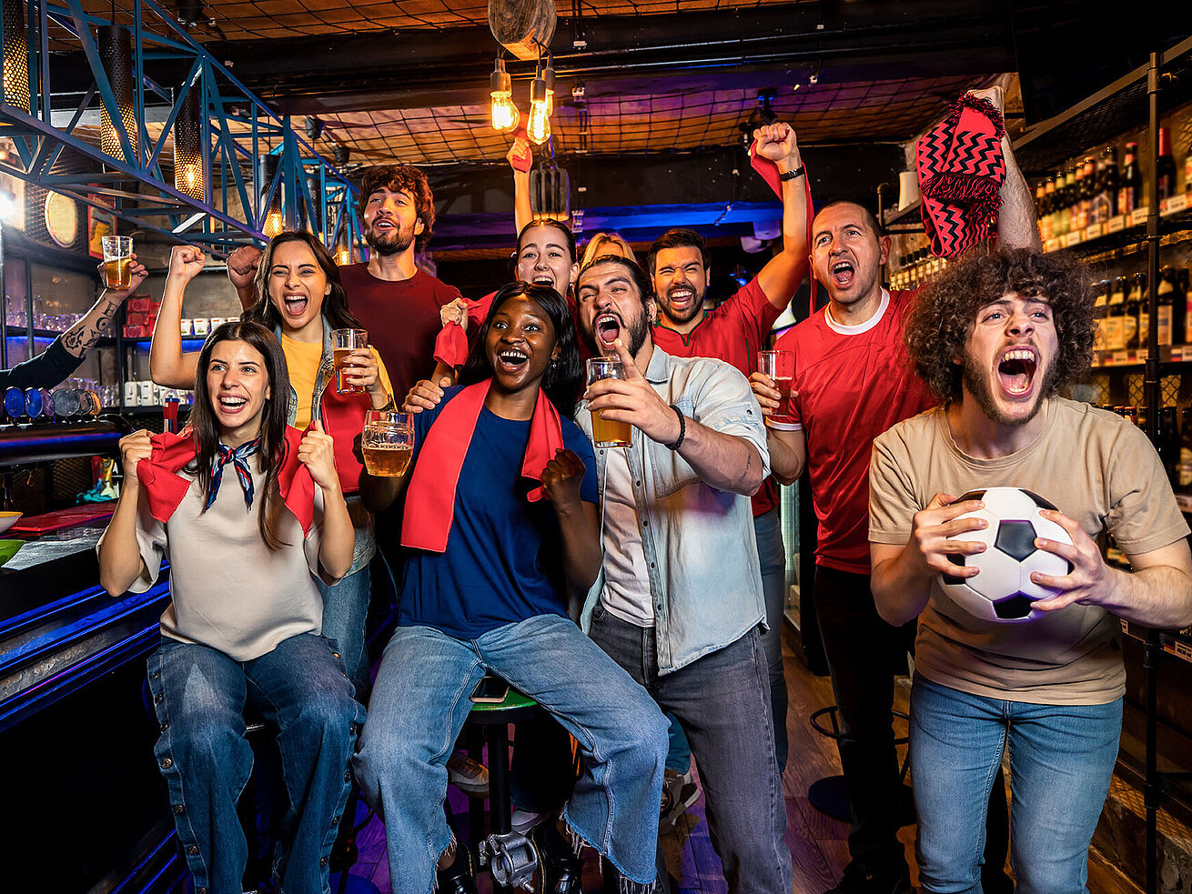 Viele Leute jubeln zusammen in einer Bar. Sie tragen Fußball-Accessoires