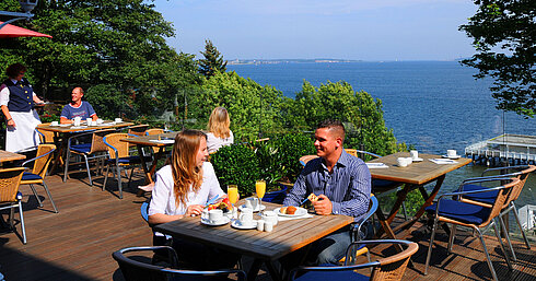 Panorama Terrasse im Maritim Hotel Kiel mit Gästen beim Frühstück, Blick auf die Förde, sonniges Wetter und entspanntes Ambiente.