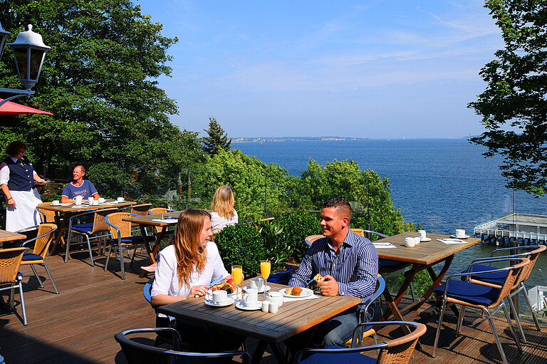 Panorama Terrasse im Maritim Hotel Kiel mit Gästen beim Frühstück, Blick auf die Förde, sonniges Wetter und entspanntes Ambiente.