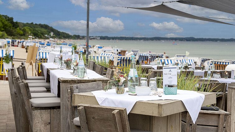 Rustikale Holztische in einer Strandlounge, gedeckt mit Wasserflaschen und Gläsern, im Hintergrund Strandkörbe und das Meer.