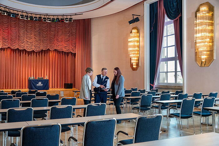 Saal Waldeck im Maritim Hotel Bad Wildungen mit Reihenbestuhlung und drei Personen im Gespräch vor der Bühne.