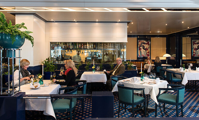 Zwei Gäste genießen ihr Mittagessen im Restaurant des Maritim Magdeburg, mit Blick auf die Bar und die Küche im Hintergrund.