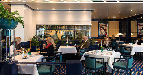 Zwei Gäste genießen ihr Mittagessen im Restaurant des Maritim Magdeburg, mit Blick auf die Bar und die Küche im Hintergrund.