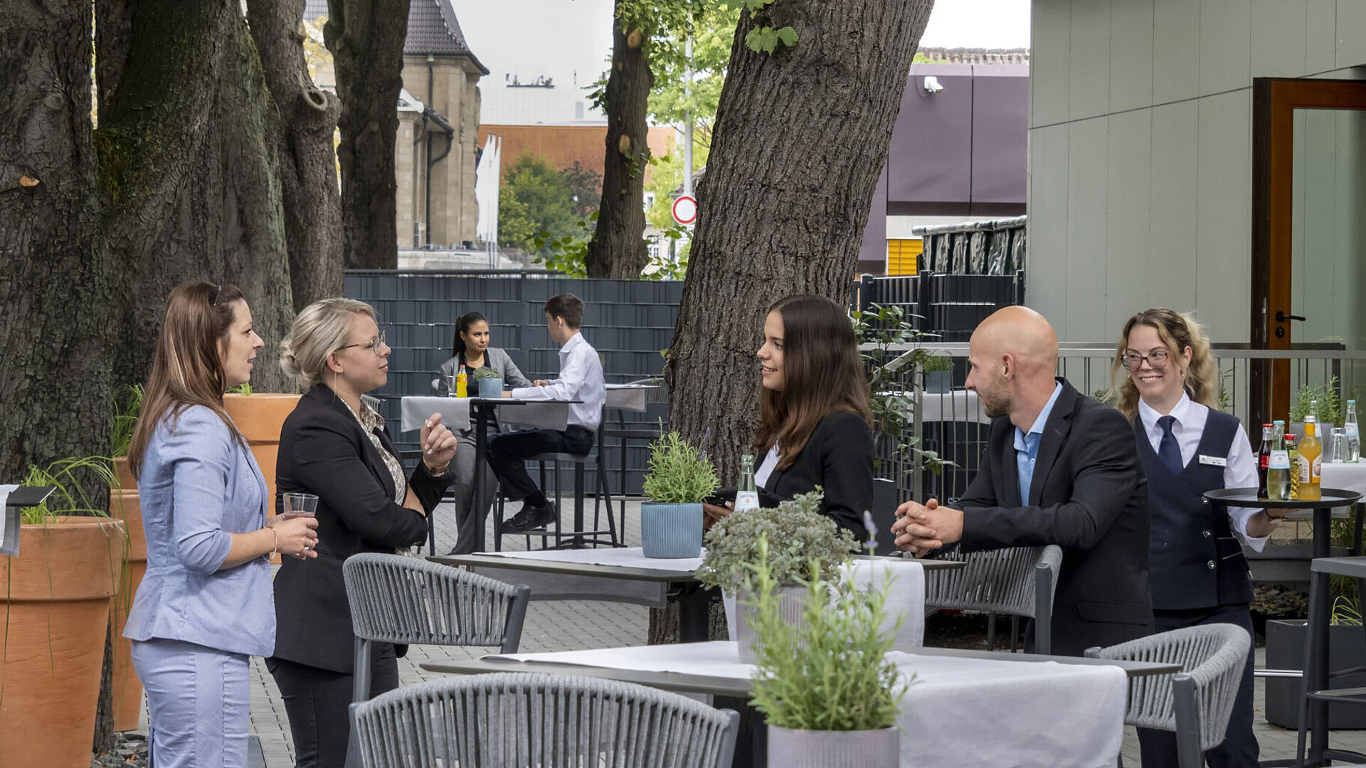 Moderne Terrasse im Maritim Hotel Darmstadt mit Gästen, die entspannen, Bäumen im Hintergrund und stilvollen grauen Möbeln.