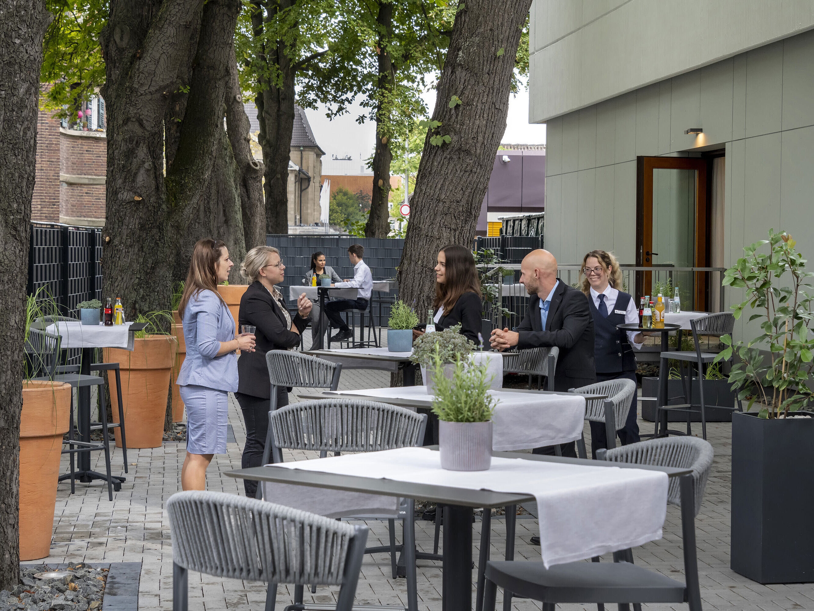 Moderne Terrasse im Maritim Hotel Darmstadt mit Gästen, die entspannen, Bäumen im Hintergrund und stilvollen grauen Möbeln.