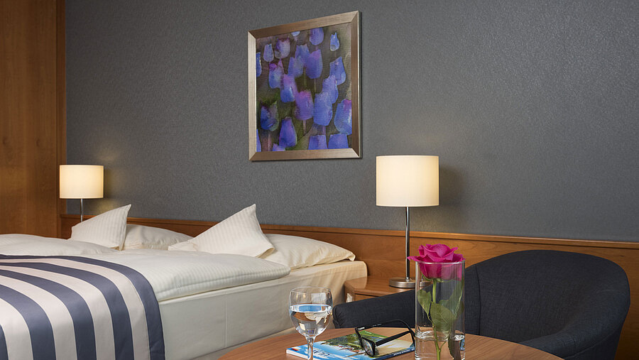 Detail im Maritim Hotel Ulm: Tisch mit Rose, Wasser, Brille und Reisebroschüre vor Doppelbett