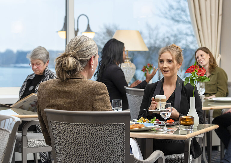 Gäste im Restaurant Bellevue im Maritim Hotel Kiel, entspannte Gespräche bei Getränken und Speisen mit Blick auf das Meer.