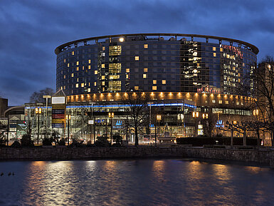 Maritim Hotel Frankfurt bei Nacht mit beleuchteter Glasfassade und Spiegelung im ruhigen Teich davor.