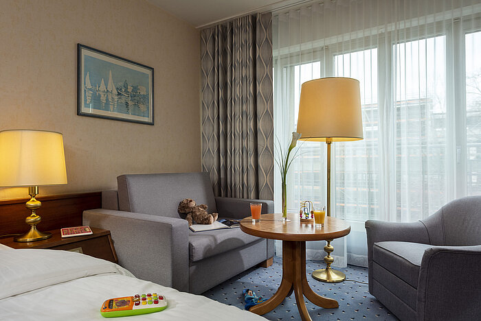 Familienzimmer im Maritim Hotel Köln mit Spielzeug auf dem Bett und Tisch, Sitzecke und großen Fenstern mit Vorhängen.