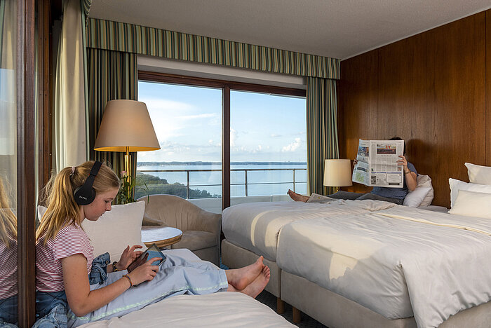 Familienzimmer mit Parkseite im Maritim Hotel Timmendorfer Strand, ein Kind mit Tablet und eine Person mit Zeitung, mit Meerblick.