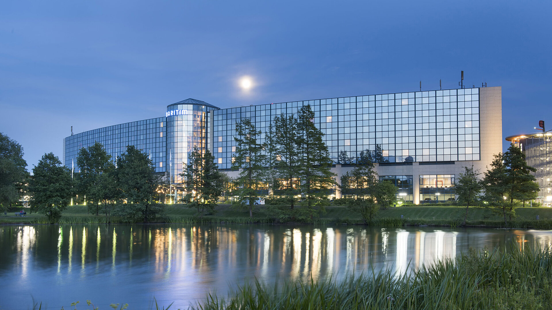Beleuchtetes Maritim Hotel Hannover mit Glasfassade und Seespiegelung in der Abenddämmerung.