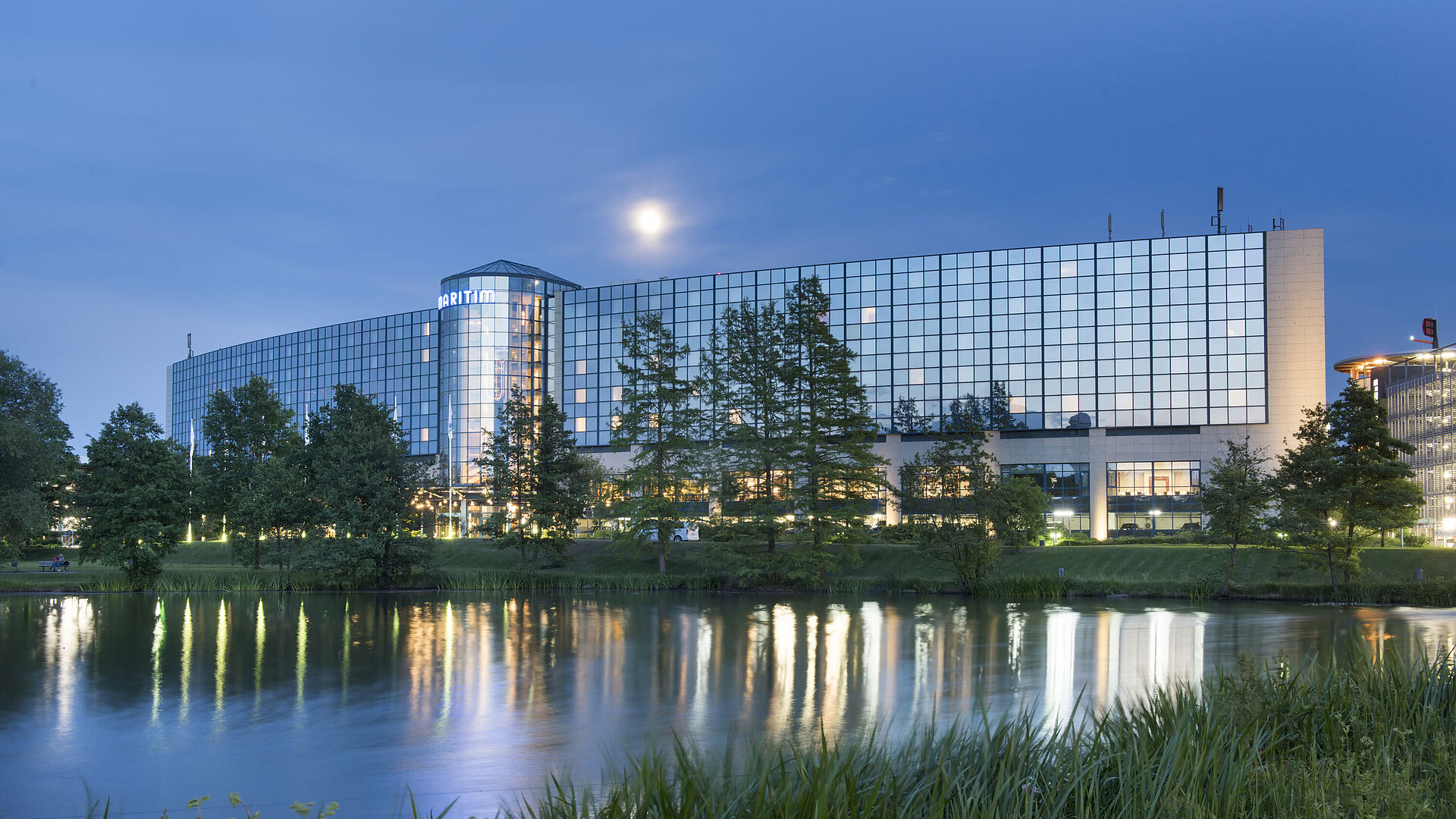 Beleuchtetes Maritim Hotel Hannover mit Glasfassade und Seespiegelung in der Abenddämmerung.