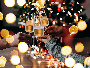 © Gregory Lee - AdobeStock.com Drei Personen stoßen mit Sektgläsern vor einem festlich beleuchteten Weihnachtsbaum an