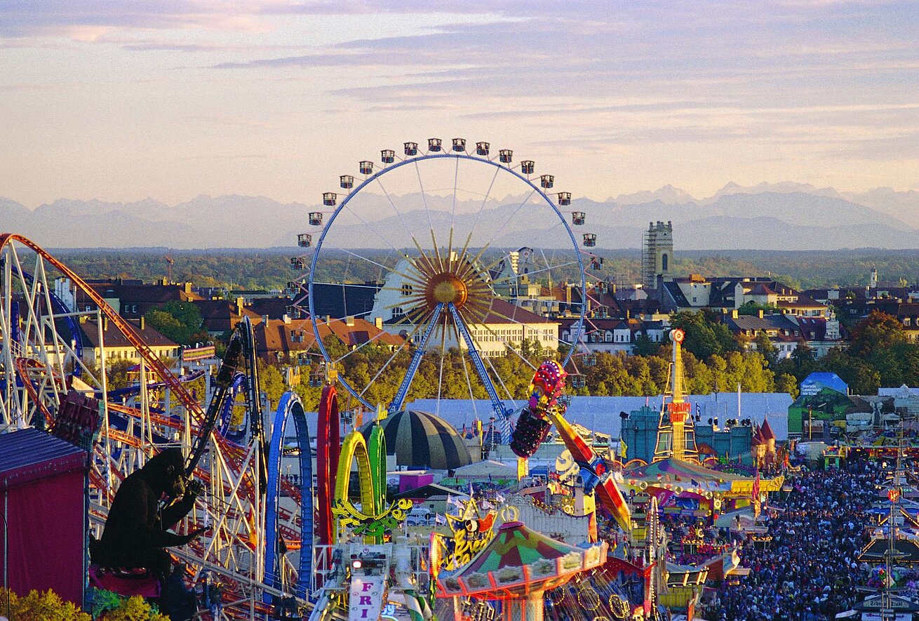 Blick über das Oktoberfest in München: Riesenrad und Fahrgeschäfte vor der Stadtsilhouette und Alpenkulisse.