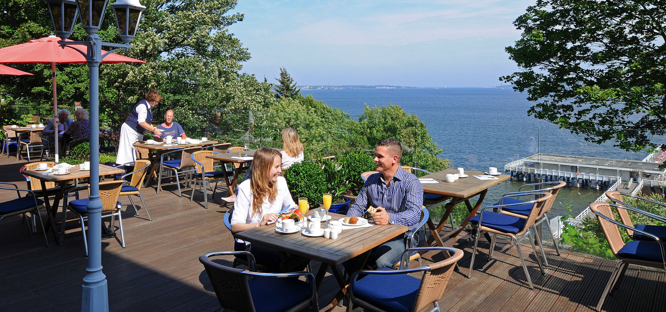 Panorama Terrasse im Maritim Hotel Kiel mit Gästen beim Frühstück, sonniges Wetter und Blick auf die Förde im Hintergrund.