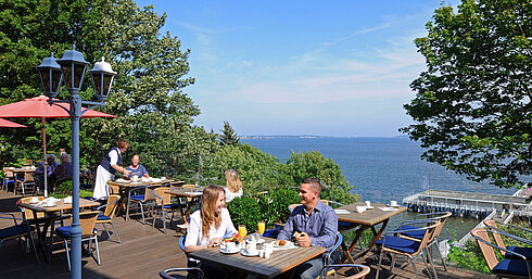 Panorama Terrasse im Maritim Hotel Kiel mit Gästen beim Frühstück, sonniges Wetter und Blick auf die Förde im Hintergrund.