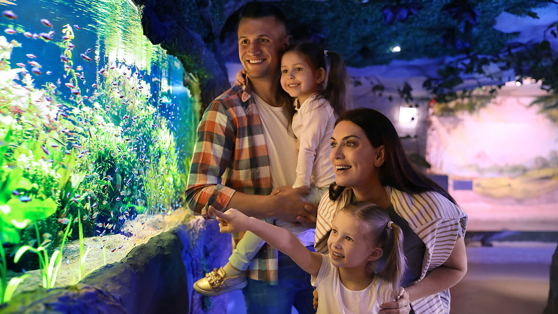 Familie betrachtet gemeinsam ein großes Aquarium, Kinder zeigen begeistert auf bunte Fische.