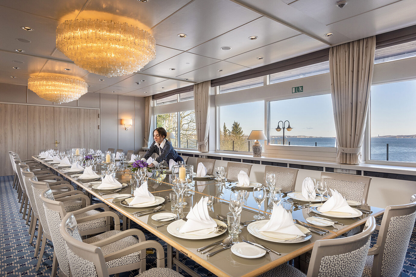 Salon Oslo im Maritim Hotel Kiel, langer festlich gedeckter Tisch mit Meerblick, Kronleuchter, Personal richtet den Raum.