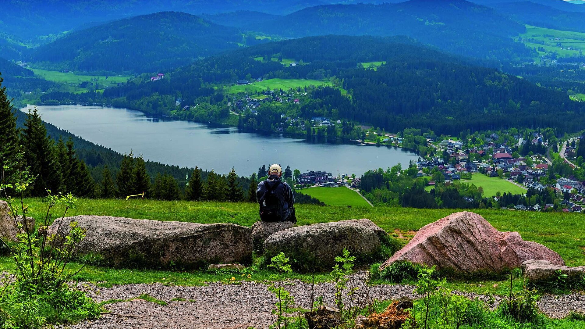 Wanderer mit Rucksack sitzt auf Felsen und schaut auf den Titisee und das Maritim Hotel im Schwarzwald