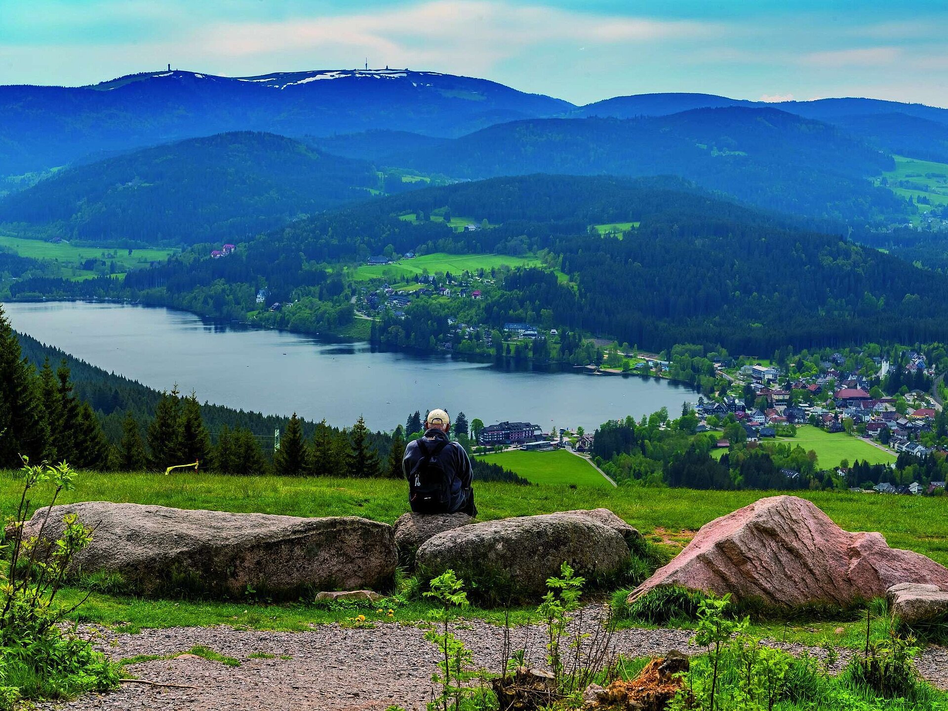 Wanderer mit Rucksack sitzt auf Felsen und schaut auf den Titisee und das Maritim Hotel im Schwarzwald
