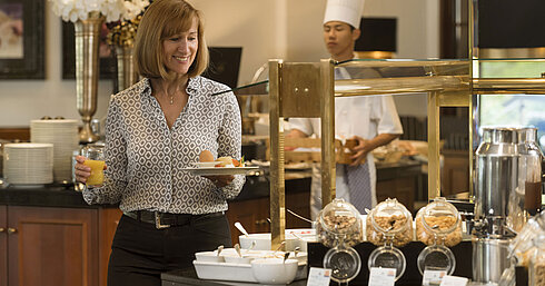 Lächelnde Hotelgast holt frische Speisen am Frühstücksbuffet im Maritim Hotel Bad Homburg, Koch im Hintergrund.