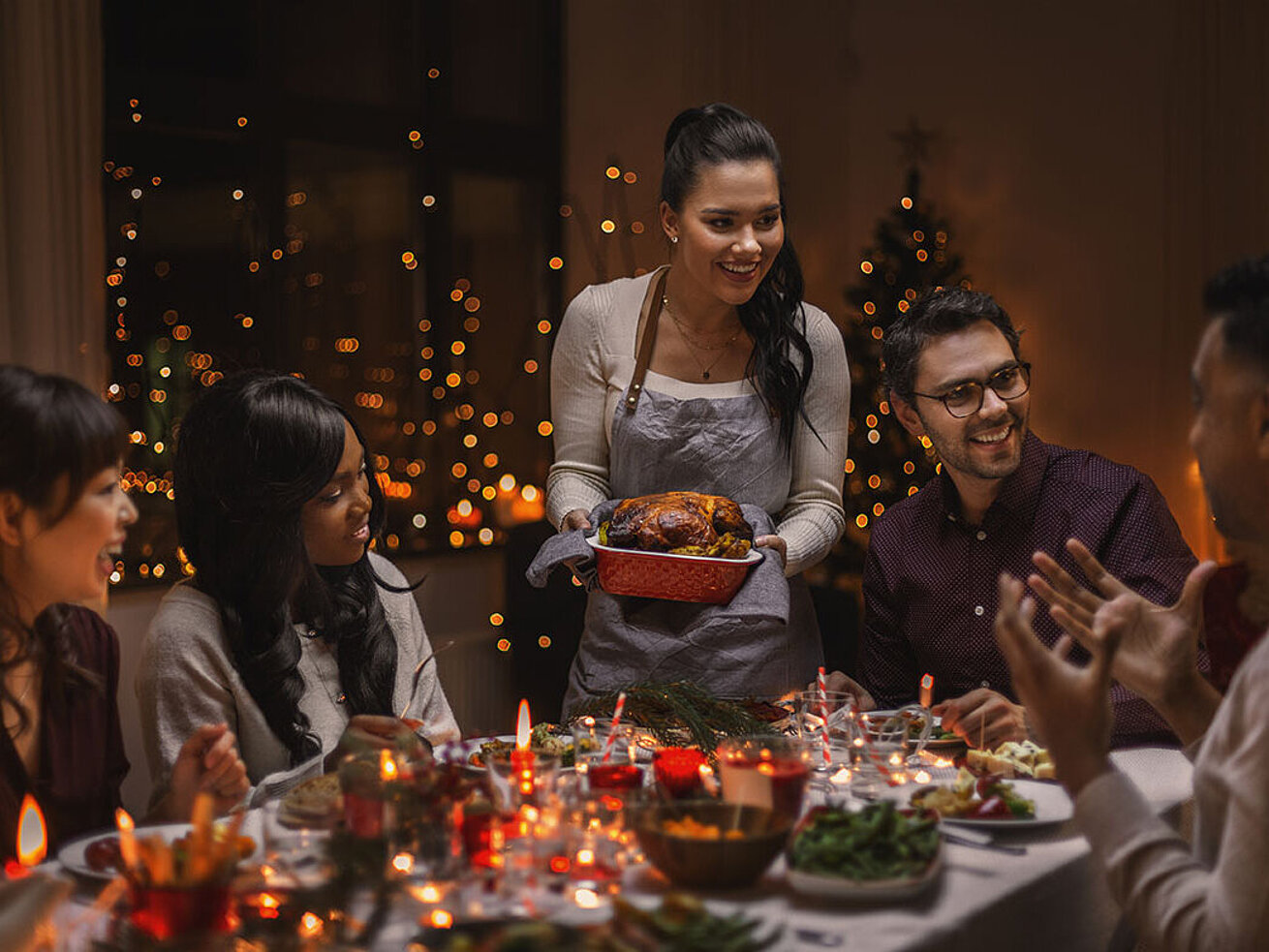 © Syda Productions - AdobeStock.com Festlich gedeckter Tisch mit Freunden beim Weihnachtsessen und einer Frau, die den Braten serviert.