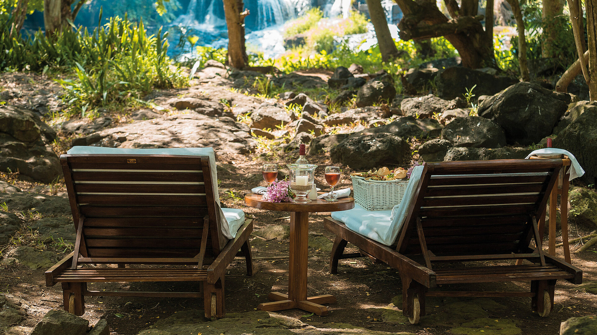 Zwei Liegestühle mit Tisch im Grünen vor Wasserfall für entspannte Auszeit