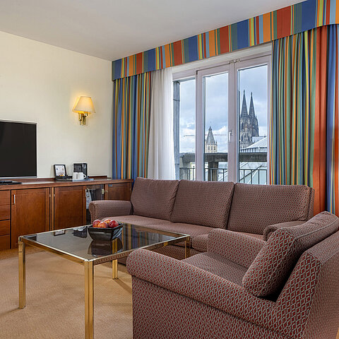 Wohnzimmer der Senior Suite im Maritim Hotel Köln mit Sofa, TV und Blick auf den Kölner Dom durch die Fenster.