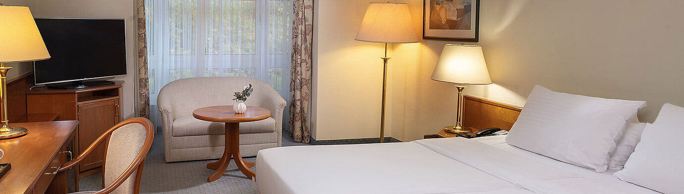 Classic Doppelzimmer mit Doppelbett, Schreibtisch, TV, Sitzbereich und großer Fensterfront in heller Einrichtung.